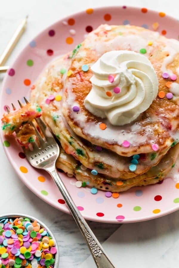 3 birthday cake funfetti pancakes on pink polka dot plate with whipped cream and vanilla icing on top.
