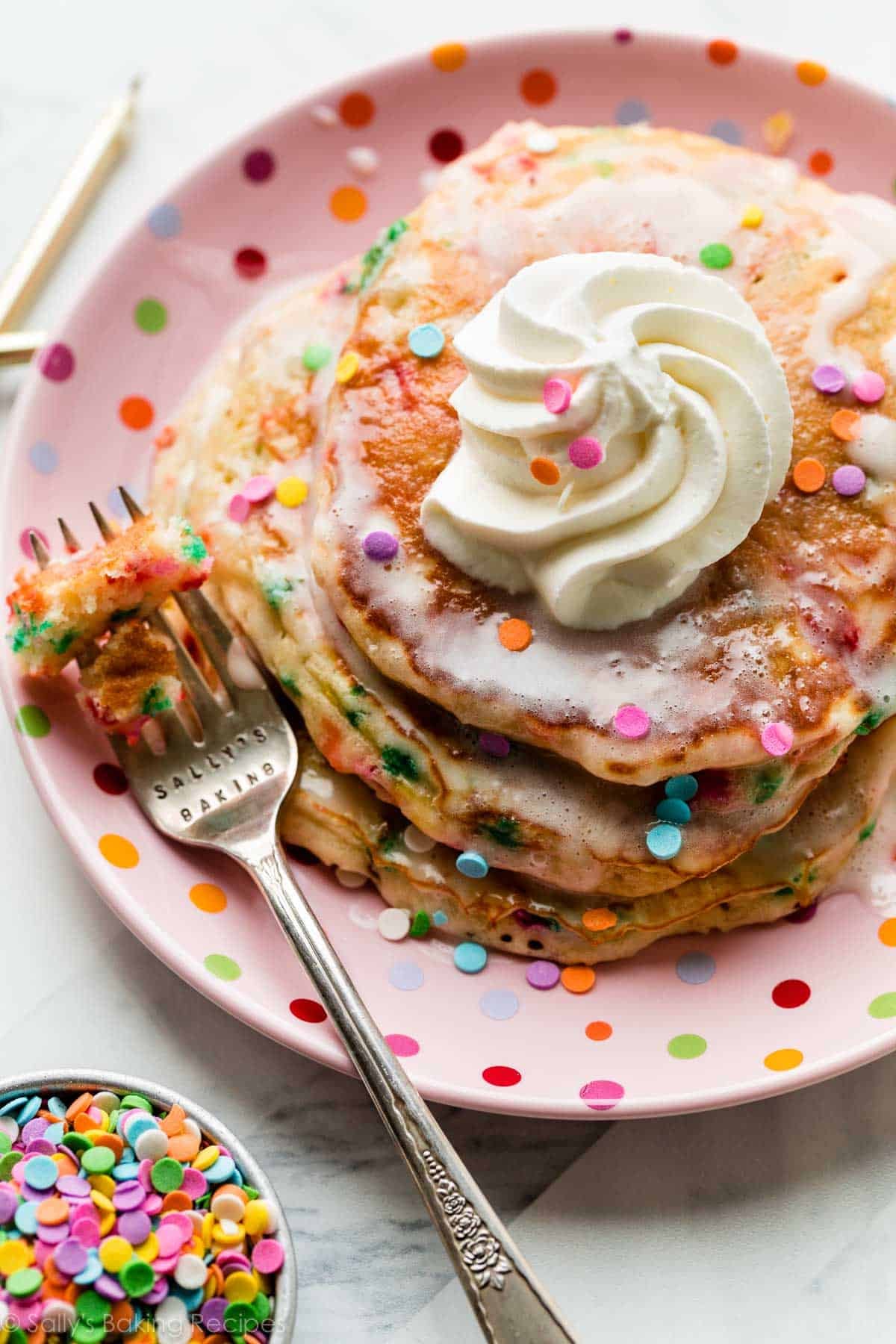 3 birthday cake funfetti pancakes on pink polka dot plate with whipped cream and vanilla icing on top.