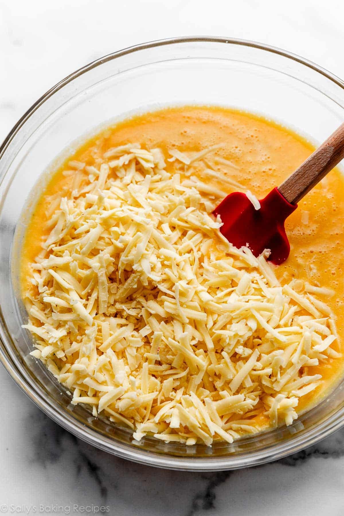 shredded gruyรจre cheese sitting on top of orange sauce in glass bowl with red spatula.