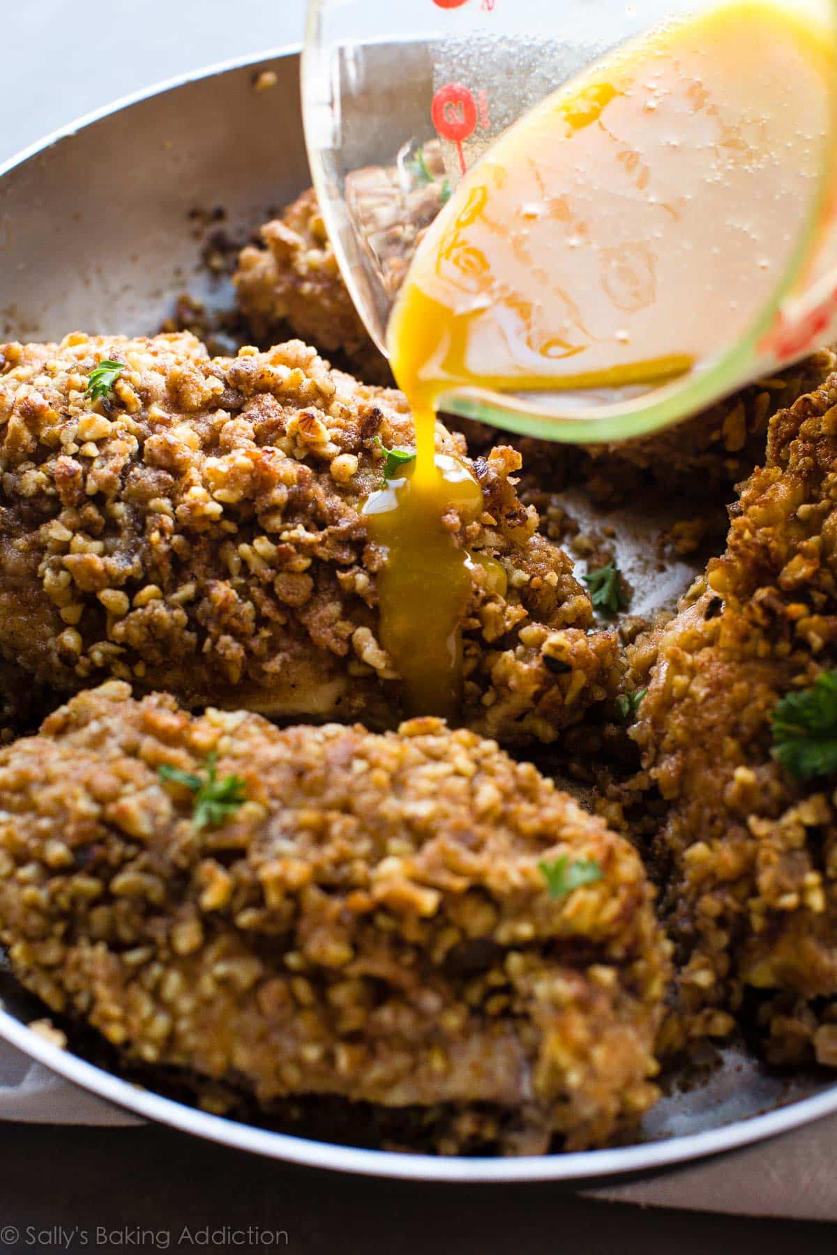 pouring honey mustard glaze onto cooked walnut crusted chicken breasts in a saucepan