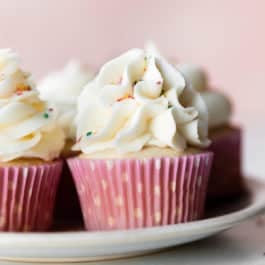 cupcakes on a plate topped with vanilla buttercream and sprinkles with pink polka dot cupcake wrappers