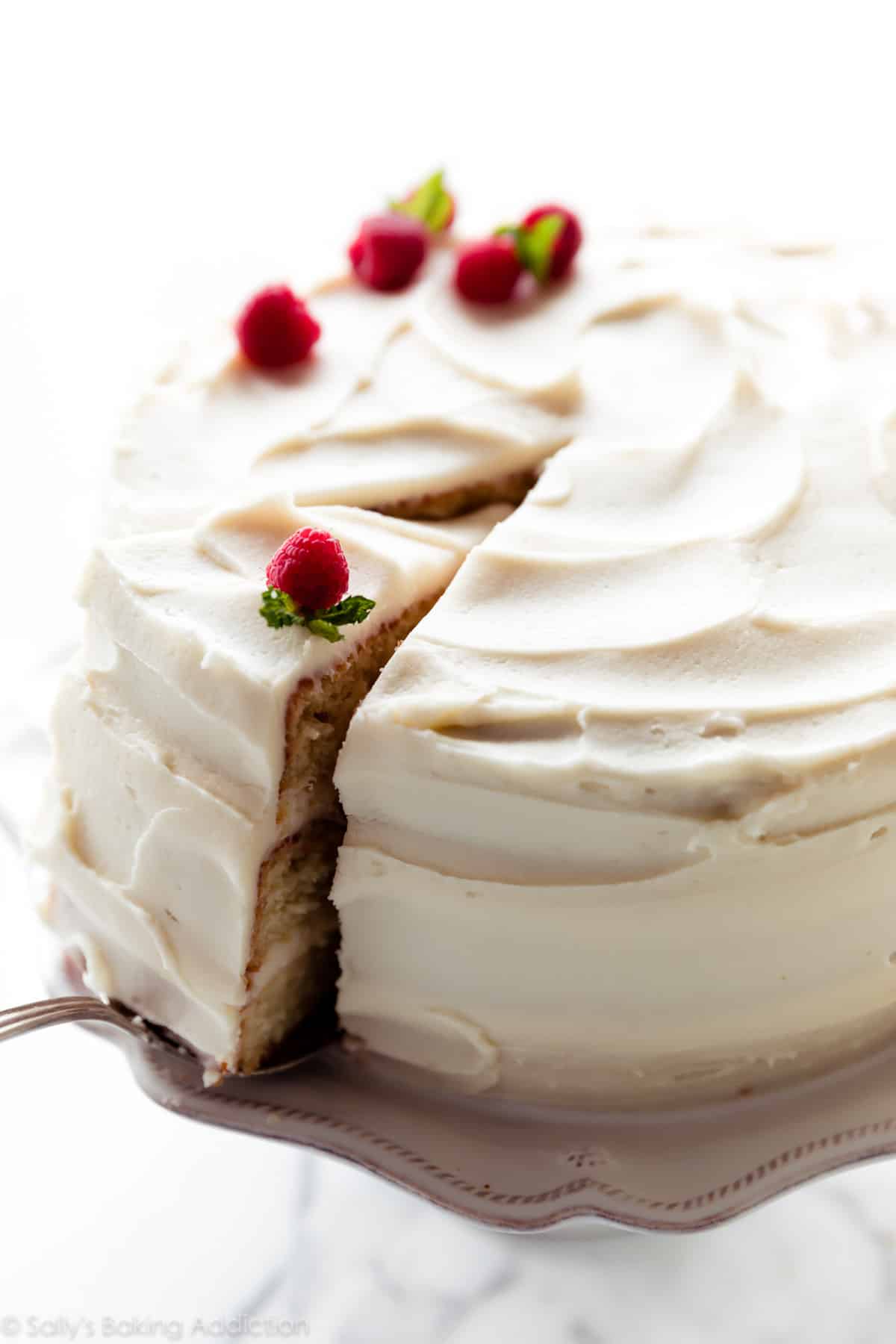 slice of vanilla cake being served from a vanilla cake with vanilla frosting on white cake stand